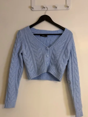 Dam Ljusblå croppad kofta med knappar - Supersöt ljusblå croppad kofta från Fbsister med stickat mönster och tre knappar framtill. Koftan har v-ringning, långa ärmar och ribbade muddar. Perfekt för lager på lager och ger en chill vibe till din outfit. Använd en gång och är som ny.