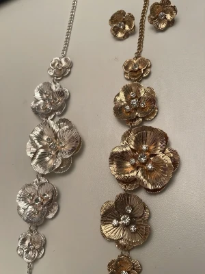 Blommig metallhalsband i silver och guld - Unik halsband med stora metallblommor i både silver och guld. Blommorna har glittrande stenar i mitten och är länkade i ett statement-mönster. Västen har en kedja som axelband och ger en riktigt cool och iögonfallande look., 150kr för den guld och 125 för silver