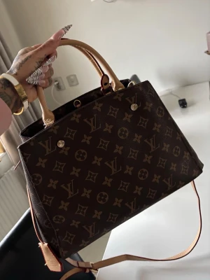 Brun handväska - Säljer en snygg handväska brunt med ikoniskt monogrammönster. Väskan har ljusa handtag, röd insida och praktisk dragkedja. Perfekt storlek för att få plats med allt du behöver och riktigt lyxig känsla.