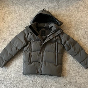 Grå Canada Goose - Grå pufferjacka från Canada Goose, modell Wyndham Parka. Jackan har huva med päls som kan sättas på. Jackan är storlek L men passar som M. Är även öppen till att byta mot en annan jacka.