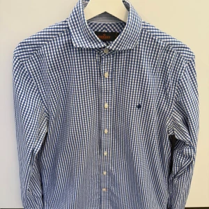 Blårutig skjorta från Morris stl 39 - Klassisk blårutig skjorta från Morris med vit botten och blå rutor. Skjortan har lång ärm, knappar framtill och en broderad logga på bröstet. Tillverkad i mjuk bomull och har en snygg krage som ger en clean look.