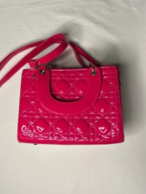 Rosa quiltad handväska med axelrem - Snygg liten handväska i glansigt rosa syntetmaterial med quiltad design och guldfärgade metalldetaljer. Väskan har både handtag och justerbar axelrem samt dragkedja upptill. Insidan är fodrad med blommigt tyg. Perfekt för dig som gillar färg och vill sticka ut.