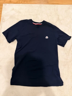 Mörkblå Moncler t-shirt med logga M/L - Snygg mörkblå t-shirt från Moncler med klassisk rund hals och korta ärmar. På bröstet sitter Monclers ikoniska logotyp i vitt och rött. T-shirten är gjord i mjuk bomull och har en diskret röd, vit och blå detalj på insidan av halsen.