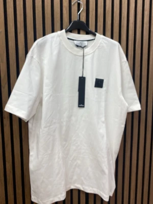 Vit t-shirt  - Vit t-shirt med klassisk rund hals och svart logopatch på bröstet. Oversized passform och tillverkad i mjuk bomull. Perfekt för en clean och stilren look.