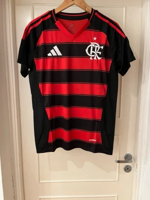 Flamengo home kit 25/26 X Jorginho 21 - Säljer en officiell Flamengo fotbollströja från Adidas med Jorginho och nummer 21 på ryggen. Tröjan har röd och svart randigt mönster, vita detaljer och v-ringad krage. Materialet är lätt polyester och perfekt för fotboll eller att representera ditt lag.