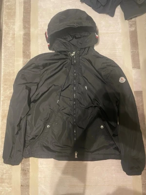 Svart Moncler vindjacka med huva - Säljer en svart vindjacka från Moncler med huva och snyggt band i rött, vitt och blått. Jackan har dragkedja framtill, två fickor med tryckknappar och Moncler-logga på ärmen. Tillverkad i polyester och har justerbar dragsko vid huvan.