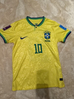 Brasilien Neymar Jr 10 Nike fotbollströja - Säljer en officiell Brasilien landslagströja från Nike med Neymar Jr och nummer 10 på ryggen. Tröjan är gul med gröna och blå detaljer, har ett subtilt mönster och är tillverkad i Dri-Fit polyester. Perfekt för fotbollsfans som vill ha något extra.