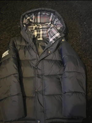 Svart pufferjacka från Burberry - Svart pufferjacka från Burberry med avtagbara ärmar så den kan användas som väst. Jackan har huva med klassiskt rutigt Burberry-foder i grått, vitt och rött. Stängs med både dragkedja och knappar. Perfekt för kalla dagar och har en riktigt schysst look.