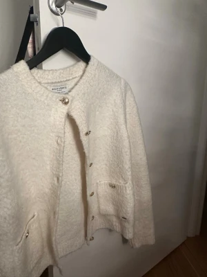 Vit kofta från Holly & Whyte - Mysig vit kofta från Holly & Whyte med rund hals, dekorativa guldknappar och två fickor framtill. Koftan är i ett fluffigt, stickat material som känns både trendigt och klassiskt. Perfekt att slänga över en topp för extra värme.