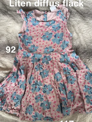 Blommig rosa barnklänning - Supersöt rosa barnklänning med stora blå blommor och små rosa hjärtan. Klänningen har volangdetaljer vid axlarna och en rund halsringning. Perfekt för sommarens alla äventyr!🌸 En diffus fläck. Storlek 92