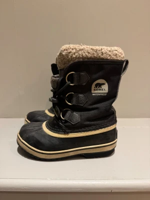 Sorel svarta vinterboots med snörning 31 - Säljer ett par klassiska svarta vinterboots från Sorel. Boots i slitstark textil och skinn, vattentäta och med grov sula. Insidan har mjukt teddyfoder med ull sula, dessa går att ta ut och och tvätta separat.   Storlek 31