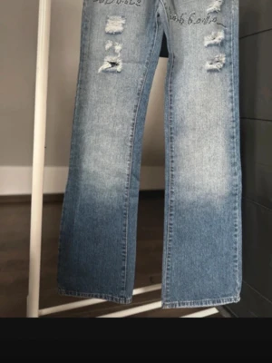 Dolce & Gabbana ljusblå jeans med strass - Unika ljusblå jeans från Dolce & Gabbana med strassdetaljer på fickan och texten 'Dolce & Gabbana' i strass framtill. Jeansen har slitna partier och raka ben med en snygg faded effekt. Perfekta för dig som vill sticka ut med coola detaljer och exklusiv känsla.