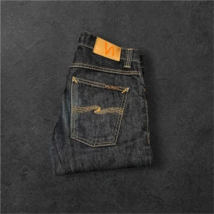 Mörkblå jeans från Nudie Jeans, W29 L32 - Snygga mörkblå jeans från Nudie Jeans med klassiska orange sömmar och logga på bakfickan. Modellen har raka ben och normal passform. Tillverkade i slitstarkt jeanstyg av bomull. Perfekta för dig som gillar stilrena och tidlösa jeans.