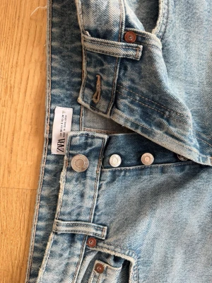 Ljusblå raka jeans från Zara - Säljer ett par ljusblå jeans från Zara med klassisk femficksdesign och knappar i silver. Jeansen har raka ben och en normal passform, med fransad kant längst ner för en cool touch. Materialet är jeans och färgen är snyggt tvättad för en avslappnad vibe.🫶🏻🤩