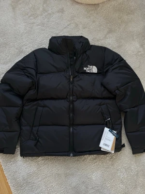 Svart pufferjacka från The North Face - Säljer en svart pufferjacka från The North Face i storlek S. Jackan har hög krage, dragkedja framtill och två sidofickor. Klassisk logga på bröstet och ryggen. Perfekt för kalla dagar och riktigt snygg streetstil.