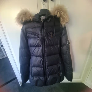 Hollies Stockholm Mörk blå pufferjacka med äktakpäls och bälte - Snygg svart pufferjacka med fluffig äktapäls på huvan och markerad midja med bälte. Jackan har flera dragkedjefickor, quiltad design och långa ärmar. Perfekt för kalla dagar när du vill hålla stilen och värmen.