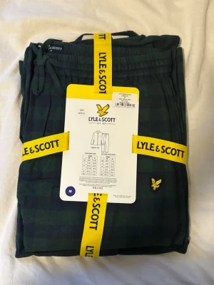 Grönrutig pyjamas från Lyle & Scott - Mysig grönrutig pyjamas från Lyle & Scott med klassisk skjortkrage och knappstängning. Setet har en bröstficka med den ikoniska gula loggan och byxor med dragsko i midjan. Perfekt för chillkvällar hemma.