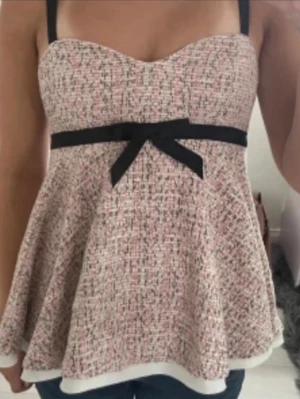 Söt blus med rosett och mönster - Supersöt blus med axelband och utställd passform. Toppens tyg har ett diskret rutigt mönster i rosa, svart och vitt. Under bysten sitter ett svart band med en stor rosett framtill. Perfekt för dig som gillar feminina detaljer.