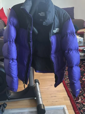 Lila och svart pufferjacka The North Face - Säljer en riktigt fet pufferjacka från The North Face i lila och svart. Jackan har hög krage, dragkedja framtill och den klassiska loggan på bröstet. Perfekt för kalla dagar när du vill hålla stilen och värmen. Passar dig som gillar streetwear och vill sticka ut.