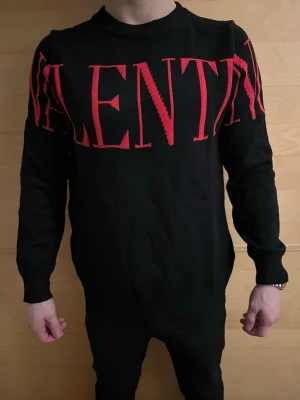 Svart Valentino tröja - Säljer en svart stickad tröja från Valentino med stora röda bokstäver över bröstet. Tröjan har rund halsringning och långa ärmar. Perfekt för dig som vill ha en statement piece i garderoben.