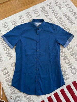 Blå kortärmad skjorta från SMOG - Säljer en blå slim fit skjorta från SMOG i storlek S. Skjortan har korta ärmar med snygg kontrastkant, klassisk krage och knäppning framtill. Tillverkad i bomull och passar perfekt till en avslappnad stil.