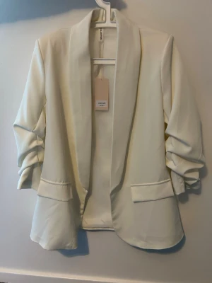 Vit kavaj med fickor från Collection - Säljer en stilren vit kavaj från Collection med snygga fickdetaljer framtill och lätt rynkade ärmar. Kavajen har en klassisk krage och är öppen framtill, vilket ger en avslappnad men ändå uppklädd look. Perfekt att slänga över en topp för en clean outfit.