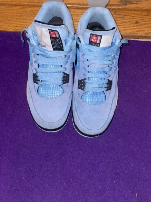 Nike Air Jordan 4 University Blue - Säljer ett par Nike Air Jordan 4 University Blue sneakers i ljusblå mocka med vita och grå detaljer samt svarta inslag. Skorna har meshpaneler, snörning och den ikoniska jorden loggan på hälen. Sulan är vit med grå stänk och svart framdel. Perfekta för dig som vill sticka ut. 