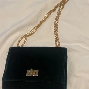 Mörkgrön sammet axelväska med kedja - Säljer en mörkgrön axelväska i mjuk sammet med guldfärgad kedjerem och snyggt vridlås i metall. Väskan har en stilren, fyrkantig form och passar perfekt när du vill addera lite lyxig känsla till din outfit.