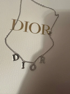 Dior halsband silver nytt - Bilden visar ett halsband från Dior i silverfärg med bokstäver och en stjärndetalj, 