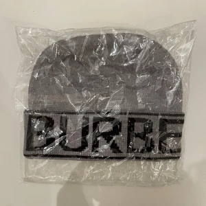 Grå burberry mössa med svart logotyp - Säljer en grå stickad mössa från Burberry med bred uppvikt kant och svart logotyptext runtom. Mössan har en klassisk form och är tillverkad i mjukt stickat material som passar perfekt för kyliga dagar.