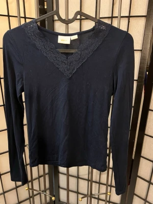 Mörkblå långärmad topp Gina Tricot - Mörkblå långärmad topp från Gina Tricot med snygg spetsdetalj runt v-ringningen. Tillverkad i mjukt och stretchigt material som består av viskos och elastan. Perfekt för dig som gillar en enkel men ändå lite extra look.