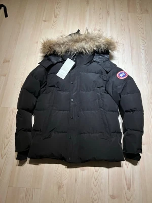 Svart jacka / Canada Goose- storlek: M - Säljer en svart jacka från Canada Goose med ikonisk patch på ärmen och stor huva med fluffig päls. Jackan har långa ärmar, dragkedja och knappar framtill samt quiltad design. Perfekt för kalla vinterdagar och riktigt snygg till streetwear. Storlek:M