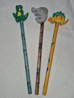 Pennor med sudd - Tre coola pennor med dinosaurietema, varje penna har ett sudd i form av en dinosaurie på toppen. Pennorna är blå, brun och gul med olika dinosauriemotiv längs hela pennkroppen. Perfekt för dig som gillar färgglada och roliga skrivsaker!