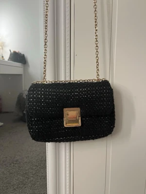Svart axelväska med kedja - Säljer en svart axelväska med guldig kedjerem och snyggt vävt material. Väskan har ett fyrkantigt guldspänne framtill som detalj. Perfekt storlek för det viktigaste och ger en stilren vibe till outfiten.