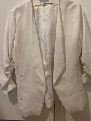 Vit kavaj från H&M, storlek 44 - Säljer en stilren vit kavaj från H&M i storlek 44. Kavajen har lång ärm, öppen front och är tillverkad i ett strukturerat tyg med diskreta metallknappar vid fickorna. Perfekt för dig som vill ha en clean och fräsch look.