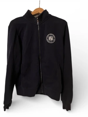 Svart ziptröja från Gant, storlek L - Klassisk svart ziptröja från Gant med hög krage och broderad logga på bröstet. Tröjan har hel dragkedja framtill, två fickor och ribbade muddar. Perfekt till lager-på-lager och enkel att matcha med allt.