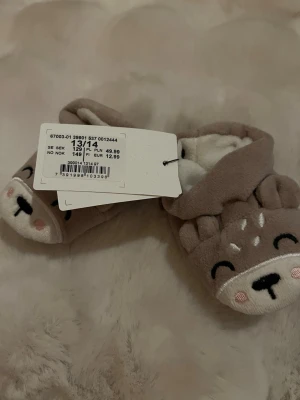 Bruna babytofflor med djurmotiv Kappahl - Supersöta bruna babytofflor från Kappahl med broderat hjortansikte och små öron framtill. Tillverkade i mjuk polyester, perfekta för att hålla små fötter varma. Insidan är vit och fluffig för extra komfort.