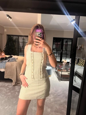 Ljus kort klänning med guldknappar - Superfin ljus kort klänning med breda axelband och fyrkantig urringning. Klänningen har ett diskret randigt mönster och dekorativa guldknappar framtill. Materialet är strukturerat och känns lite som tweed, vilket ger en lyxig känsla.