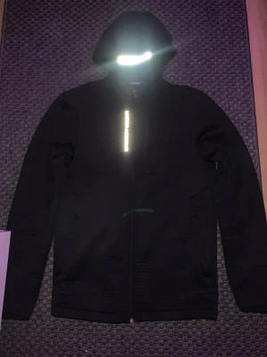 Svart vindjacka från Under Armour - Säljer en svart vindjacka från Under Armour med huva och reflekterande detaljer fram och bak. Jackan har dragkedja hela vägen och är tillverkad i ett lätt syntetmaterial. Perfekt för dig som vill ha en sportig look och gillar funktionella plagg. Helt oanvänd och fräsch!