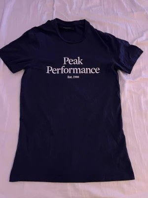 Mörkblå Peak Performance t-shirt S - Snygg mörkblå t-shirt från Peak Performance med vit logga och text på bröstet. Klassisk rund halsringning och korta ärmar. Tillverkad i mjuk bomull som är skön att ha på sig hela dagen.