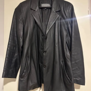 Svart skinnkavaj från Valentino- Vera Pelle  - Snygg svart kavaj i äkta skinn från Valentino. Klassisk modell med breda slag, fyra knappar framtill och två fickor. Perfekt för dig som vill ha en stilren och edgy look. Passar till många olika outfits och ger en cool vibe.