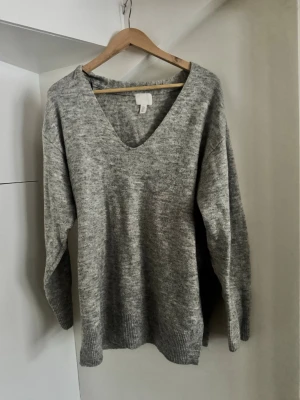 Grå stickad tröja med v-ringning H&M - Mysig grå stickad tröja från H&M med djup v-ringning och långa ärmar. Tröjan har ribbade muddar vid ärmslut och nederkant, perfekt för lager på lager under kyliga dagar. Enkel och stilren design som passar till allt.