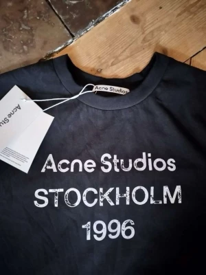 Svart långärmad topp Acne Studios - Svart långärmad topp från Acne Studios med vit text 'Acne Studios STOCKHOLM 1996' framtill. Klassisk rund halsringning och rak passform. Tillverkad i mjuk bomull, perfekt för en clean och stilren look. Pris kan diskuteras!