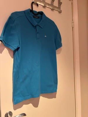 Blå pikétröja från J.Lindeberg - Säljer en klassisk blå pikétröja från J.Lindeberg i storlek S. Tröjan har korta ärmar, krage och knappar framtill samt en liten vit logga på bröstet. Perfekt till sommaren och enkel att matcha med jeans eller shorts.