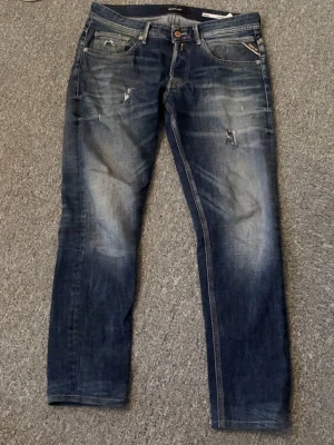 Replay blå jeans med slitningar - Säljer ett par blå Replay jeans med snygga slitningar och tvättade detaljer. Klassisk femficksmodell med raka ben och normal passform. Jeansen har läderpatch bak i midjan och tydliga Replay-detaljer. Perfekta för en avslappnad och trendig look.