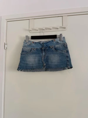 Blå jeanskjol med broderade fickor - Säljer en kort jeanskjol i ljusblå denim med råa, fransiga kanter och slitningar. Kjolen har klassiska fickor fram och bak, där bakfickorna är dekorerade med stora broderade kors i beige och rosa toner. Perfekt för dig som gillar en edgy och unik stil. De är två kjolar från början som jag gjort om o sytt o grejer. Använd fåtal gånger.