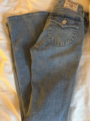 True Religion ljusblå bootcut jeans - Säljer ett par ljusblå jeans från True Religion i klassisk bootcut-modell. Jeansen har snygga kontrastsömmar, ikonisk fickdetalj med lock och knapp bak samt femficksdesign. Tillverkade i mjukt jeansmaterial med normal passform och midwaist.