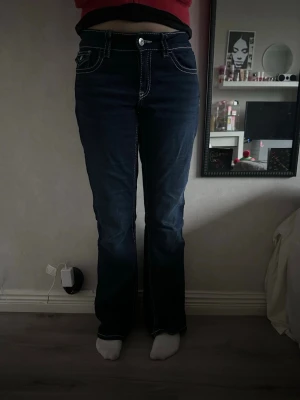 Mörkblå bootcut jeans med kontrastsömmar - Säljer ett par mörkblå jeans med tydliga vita kontrastsömmar och snygga fickdetaljer bak. Modellen har bootcut-ben och normal passform, perfekt för dig som gillar en klassisk men ändå trendig look. Jeansen har normal midja och är tillverkade i klassiskt denimtyg.