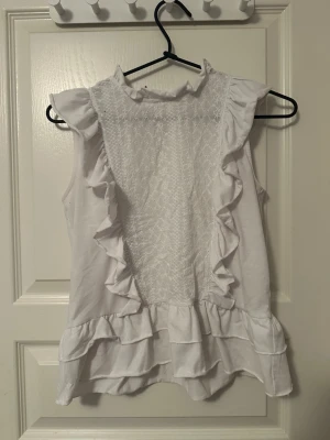 Vit ärmlös blus med volanger - Säljer en söt vit blus med volangdetaljer längs axlar och nederkant. Framsidan har ett broderat mönster och hög hals med liten volangkant. Blusen är ärmlös och har en luftig passform, perfekt till sommaren!😽Priset kan förhandlas!