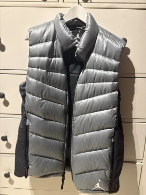 Silver dunväst från Jordan - Säljer en riktigt snygg silverfärgad dunväst från Jordan. Västen har quiltad design, hög krage och dragkedja framtill. Två fickor med dragkedja på sidorna och den klassiska Jumpman-loggan nedtill. Perfekt för lager på lager under höst och vinter. Lite större i storlek, västen är storlek M men passar också storlek L